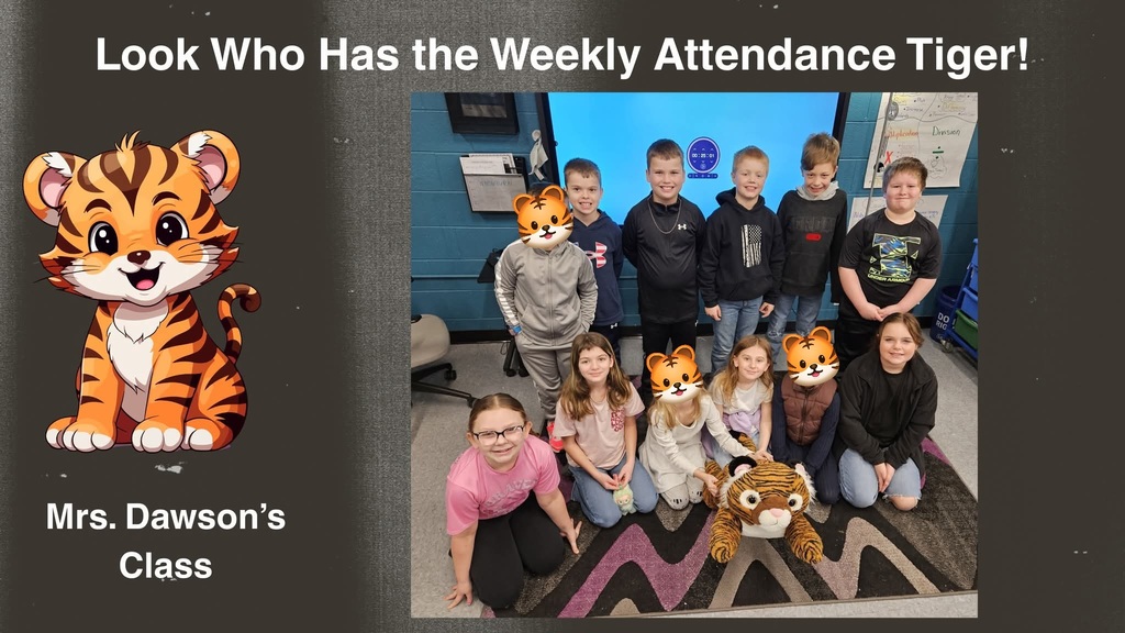 Students from Mrs. Dawson’s class are gathered together in their classroom for a group photo. Some students are sitting on a patterned rug, and others are standing behind them. A large stuffed tiger, representing the Attendance Tiger, is placed at the front of the group. Behind the students is a large interactive screen and classroom materials on the walls.