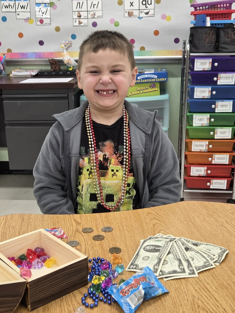Winston Duncan is seated at a classroom table wearing a dark jacket and a shirt with a colorful graphic design. The student has several bead necklaces around their neck. On the table in front of the student are an open wooden box filled with colorful plastic jewels, several coins, a few one‑dollar bills, strands of beads, and a small pack of candy. Behind the student is a classroom bulletin board with instructional cards and colorful storage bins.
