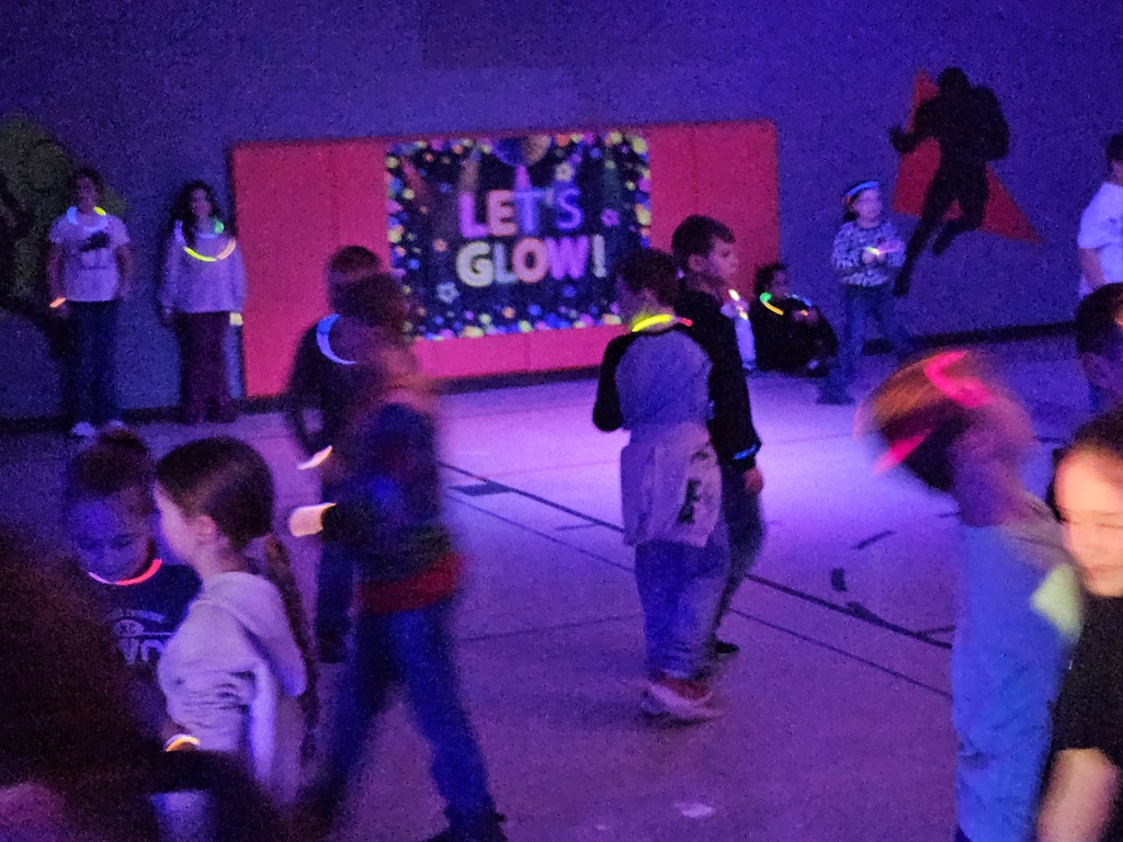 Students in a school gym participating in a glow‑themed dance party. The room is dimly lit with colorful glowing accessories such as glow sticks. A teacher is present acting as the DJ for the event. This photo accompanies an announcement about Wirt County Primary Center’s February Super Secret Day, which featured a glow dance party to boost student attendance