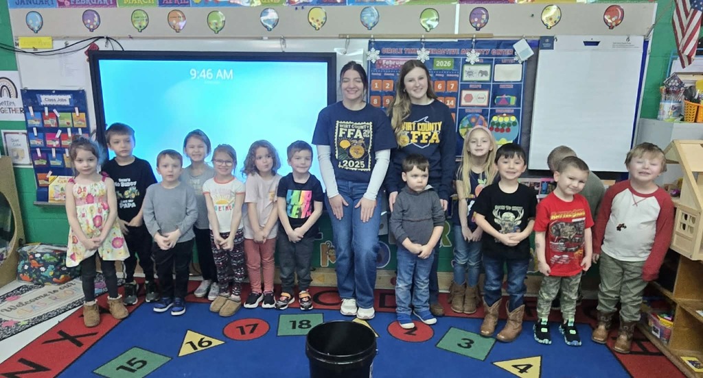 A group of young children standing in a line on a colorful rug in a classroom with two older students wearing FFA shirts standing behind them. A large digital screen is visible on the wall, along with bulletin boards and classroom materials. The children and the FFA students are posed together for a group picture.