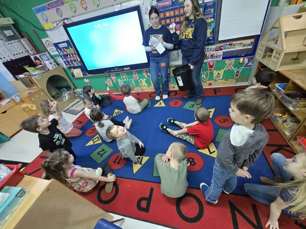A classroom scene showing a group of young children seated on a colorful rug with numbers around the border. Two older students wearing matching shirts with FFA logos stand at the front of the room holding papers and reading to the class. Behind them is a large digital screen and a wall decorated with letters and educational materials. The children are seated in a semi‑circle, looking toward the readers.