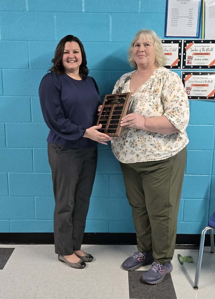 Carla Brown and Annette Easton standing in front of a bright blue painted brick wall. One holds a wooden recognition plaque with engraved nameplates while the other stands beside. On the wall behind them are classroom posters and award certificates. Both individuals are centered and facing the camera.