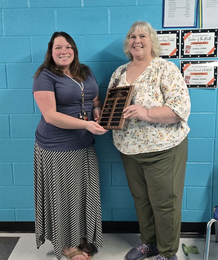 Jessica Arthur and Annette Easton standing in front of a bright blue painted brick wall. One holds a wooden recognition plaque with engraved nameplates while the other stands beside. On the wall behind them are classroom posters and award certificates. Both individuals are centered and facing the camera.