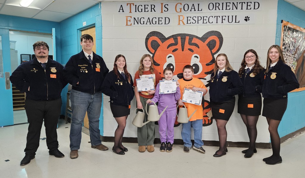 A group of older students wearing matching jackets with FFA emblems stand in a school hallway alongside three younger students holding certificates and light‑colored plastic watering cans. The group is standing in front of a large wall display featuring a tiger illustration and the words “A Tiger Is Goal Oriented, Engaged, Respectful.” The setting includes brightly painted blue walls, a bulletin board on the right, and a hallway entrance on the left. Everyone is standing in a line facing the camera while the three younger students display their awards.
