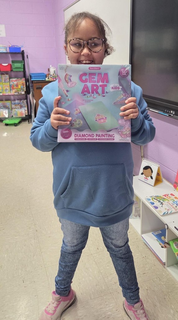 A student standing indoors holding a boxed Gem Art Diamond Painting kit in front of the torso. The kit packaging is pastel‑colored with the words “Gem Art Kids Diamond Painting” visible. The student is wearing a blue hoodie, jeans, and sneakers. The classroom setting includes shelves with books and materials, a purple wall, and part of a digital display screen on the right side of the image.