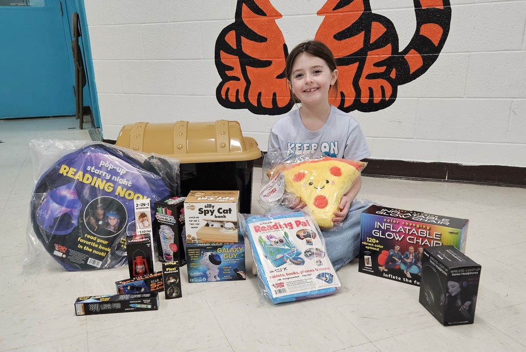 A child is seated on the floor in front of a wall painted with a large orange tiger graphic. The child is surrounded by an assortment of prizes arranged in a semicircle. Visible items include a round “Reading Nook” pop‑up tent package, a plastic treasure‑chest style toy box, a plush pizza‑slice pillow, a “Reading Pal” animal‑shaped reading cushion, a boxed inflatable glow chair, a “Silly Spy Cat” toy, a “Galaxy Gelly” set, a book light, a mini lava lamp, and other small prizes. The setting appears to be a school hallway with bright walls and tiled flooring.