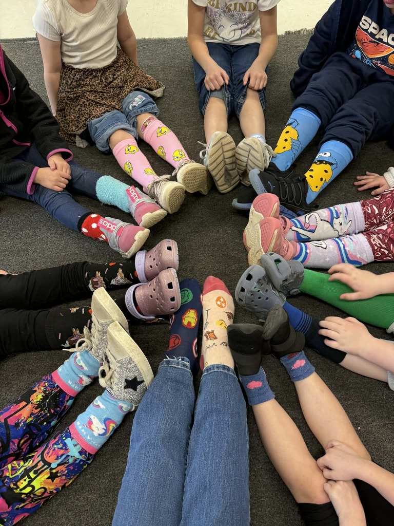A group of students sitting in a circle on a carpeted floor with their legs stretched toward the center. Each student is wearing brightly colored or patterned socks as part of a crazy‑sock activity. The socks feature a variety of designs, including cartoon characters, animals, stripes, polka dots, food themes, and vibrant abstract patterns. Some students are wearing sneakers, slip‑on shoes, or Crocs, while others have removed their shoes to show their socks. Only the lower legs, feet, and partial torsos of the students are visible.