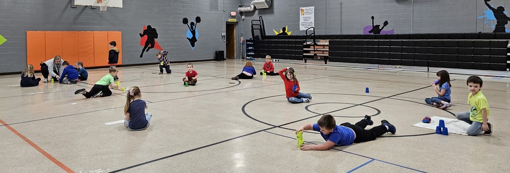 A group of elementary students from Mrs. Vannoy’s class are spaced around the gym floor, participating in an activity while wearing red and blue shirts for Reading Week. Some students are seated, and others are lying or leaning forward while interacting with plastic stacking cups. The gym has painted sports silhouettes on the walls, orange wall padding on the left, and a set of black bleachers in the background. The students are positioned in small groups or individually around the center circle markings on the gym floor.