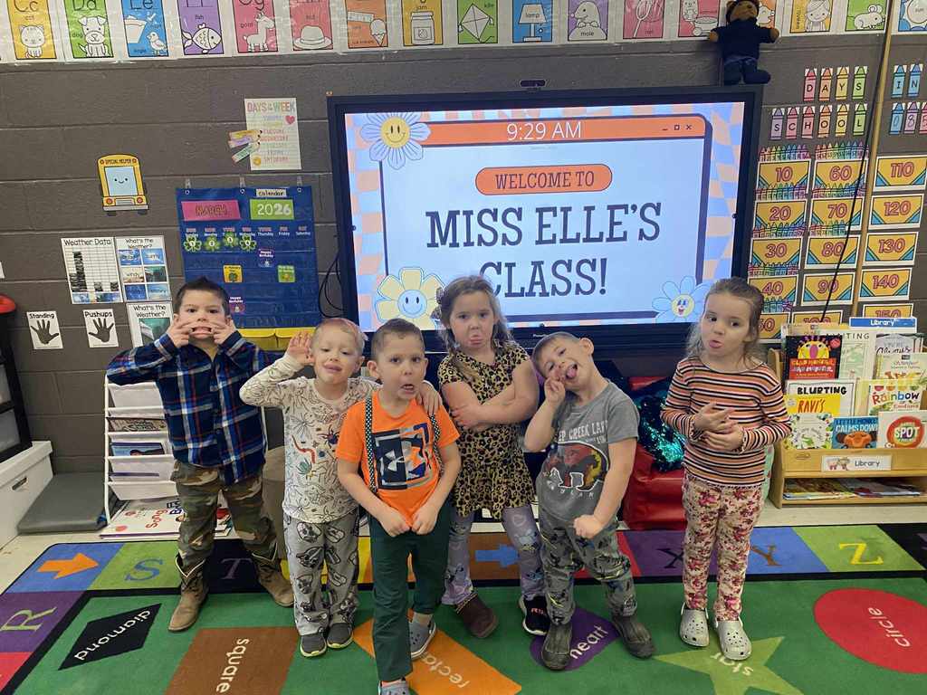 A group of seven students stand in front of an interactive board displaying a slide that reads “Welcome to Miss Elle’s Class!” The students are wearing a mix of patterned clothing, including animal prints, colorful shirts, and mismatched outfits. Classroom posters, bins, books, and a vibrant rug with letters and shapes are visible throughout the room.