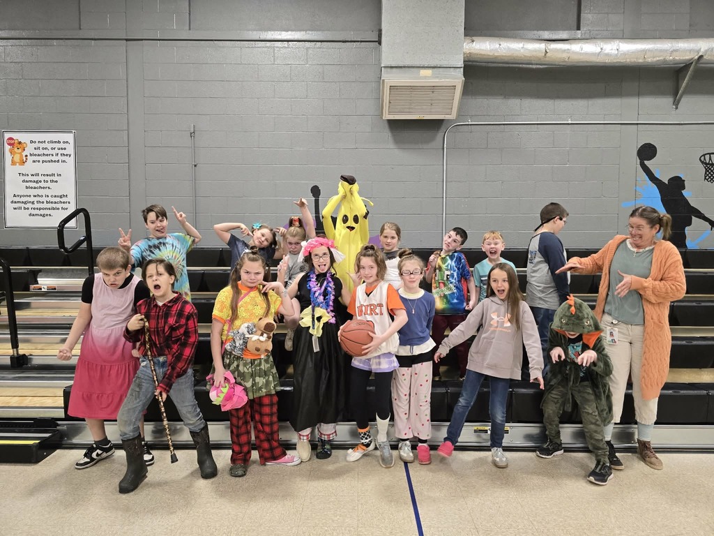 A large group of students stand in a gymnasium in front of bleachers, dressed in a wide variety of creative Wacky Wednesday outfits. Clothing includes bright tie-dye shirts, costumes, patterned pants, sports jerseys, a banana costume, and accessories such as hats, scarves, and necklaces. A basketball and a walking cane are also visible among the group. The gym walls are gray with painted sports silhouettes.