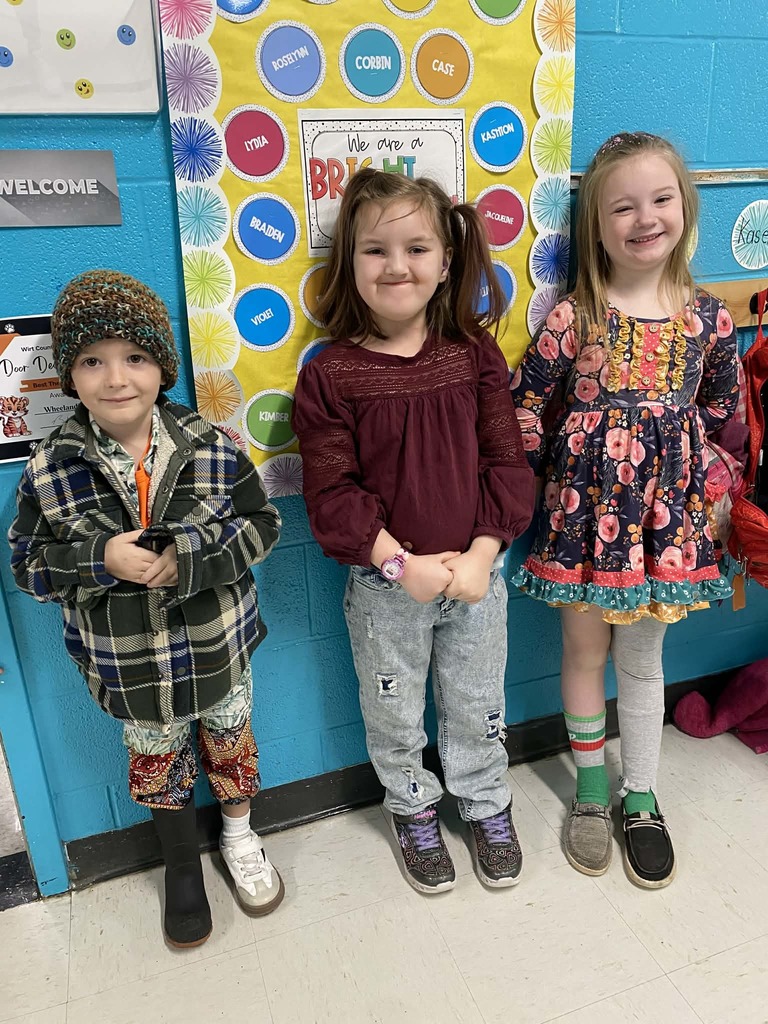 Three students stand side by side in a school hallway in front of a bright bulletin board decorated with colorful circles and the words “We Are a Bright Bunch.” The student on the left is wearing a knit hat, plaid jacket, patterned pants, and mismatched boots. The student in the middle is wearing a burgundy top and jeans with patches. The student on the right is wearing a floral patterned dress with layered colors, mismatched socks, and different shoes.