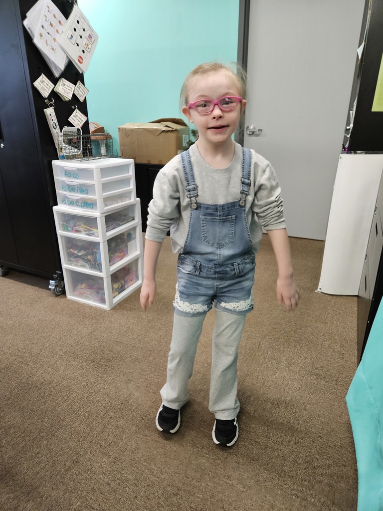 A student stands in a classroom area wearing layered clothing: denim overalls, long pants underneath, and a long‑sleeve top. The student is standing on a carpeted floor with storage drawers, a door, and various classroom materials visible in the background.
