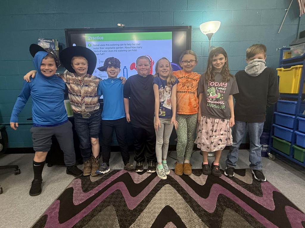 Eight students stand in a classroom in front of an interactive board displaying a slide with text and graphics. The students are wearing a variety of Wacky Wednesday outfits, including cowboy hats, bright shirts, patterned leggings, and themed graphic tees. Shelving, bins, and a lamp are visible around the room, and a zigzag-patterned rug covers part of the floor.