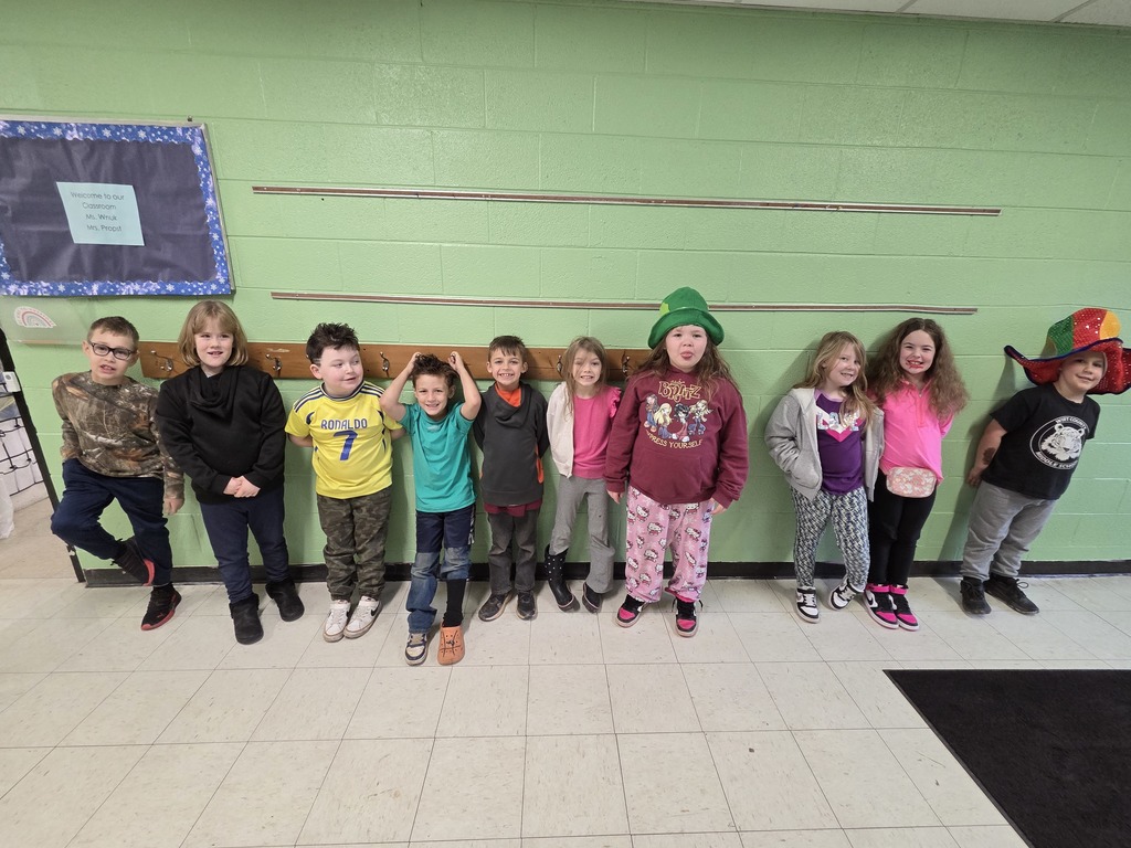 A group of students stands in a school hallway against a light green painted wall. They are lined up in a row beneath a set of metal coat hooks. The students are wearing a variety of colorful and mismatched outfits for Wacky Wednesday, including patterned pants, bright shirts, hooded sweatshirts, jeans, and a large green hat worn by one student. Another student on the far right is wearing a multicolored jester‑style hat. A bulletin board with a blue border is visible on the left side of the wall, and the floor is tiled in white and light gray.