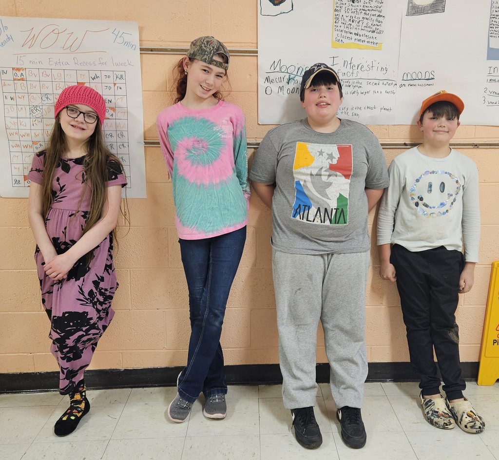 Four children stand in a line in a school hallway against a peach‑colored wall. Behind them are large classroom posters containing handwritten information and charts. Each child is wearing a different type of hat, including a knitted hat, baseball caps in different colors, and a brimmed cap. Their outfits include a dress with a bold floral pattern, a tie‑dye shirt, a graphic T‑shirt with a multicolored design, and a long‑sleeved shirt with a printed character face. The floor is tiled in light and darker shades.