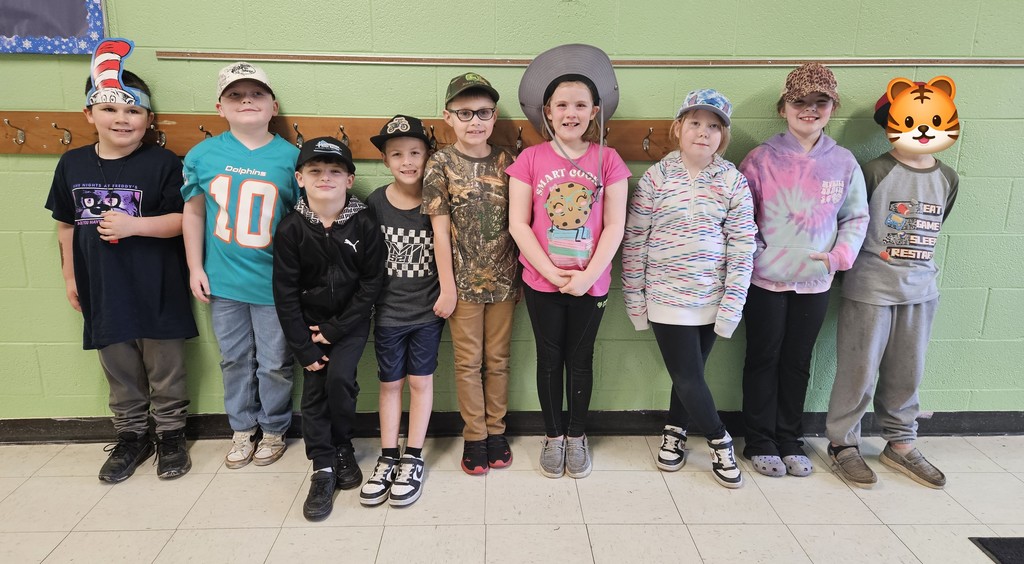 A group of ten children stand side by side in a school hallway against a light green wall. They are wearing a variety of hats and clothing for Hat Day. Visible hats include a tall striped hat, baseball caps, an oversized wide‑brimmed hat, and a headband with a small printed design. Their outfits range from graphic T‑shirts and hoodies to patterned tops and camouflage designs. The children are standing in front of a wooden coat rack mounted to the wall. The floor is tiled in light and dark squares.