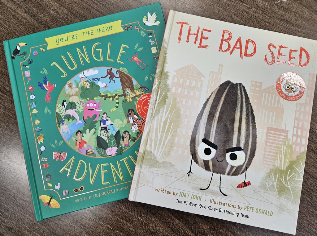 Two children’s books are shown lying side by side on a wooden surface. The book on the left has a green cover and is titled “You’re the Hero: Jungle Adventure,” featuring illustrated people, animals, plants, and insects arranged around a circular jungle scene. The book on the right has a light-colored cover and is titled “The Bad Seed,” displaying an illustrated sunflower seed character standing on a sidewalk with buildings in the background. Both books are clearly visible and placed close together.