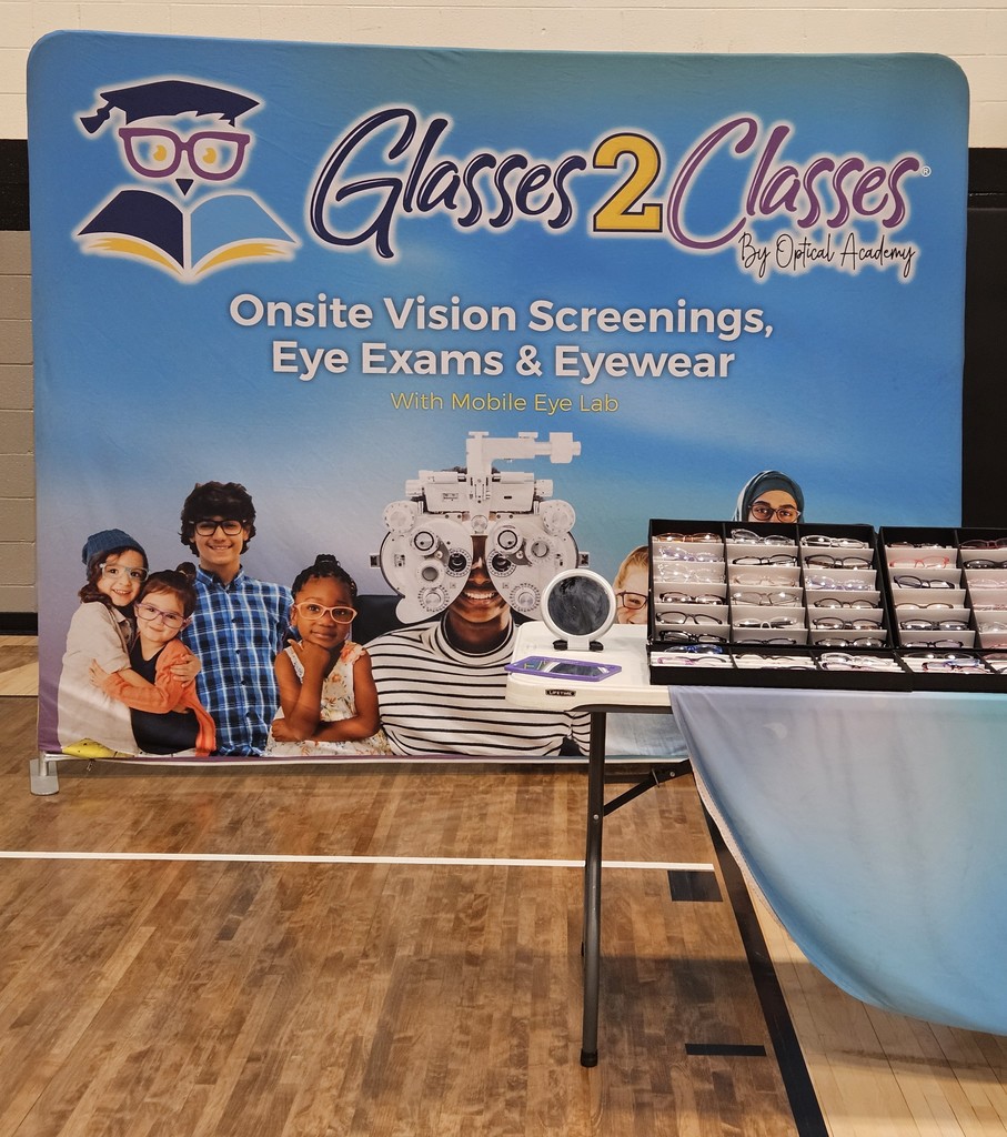 Photo taken inside a school gym showing a Glasses 2 Classes display setup. A large blue backdrop sign reads “Glasses 2 Classes by Optical Academy” with text underneath that says “Onsite Vision Screenings, Eye Exams & Eyewear” and “With Mobile Eye Lab.” The sign includes images of children wearing glasses and optical testing equipment. In front of the backdrop, a table displays rows of eyeglass frames organized in cases. The gym floor and wall padding are visible around the display.