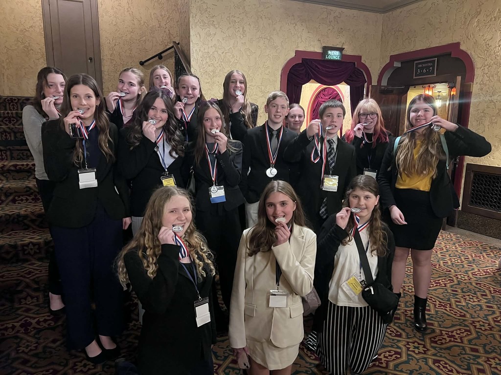 A group of smiling students in business suits standing indoors, proudly holding up their silver competition medals for the camera.