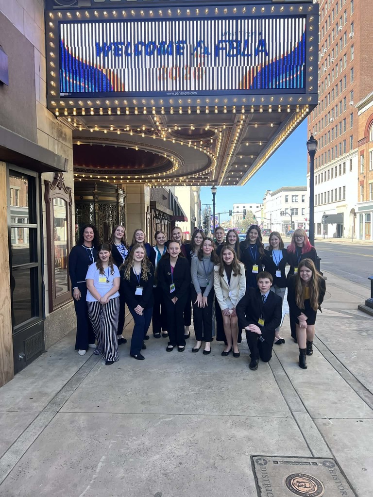 A medium shot shows a diverse group of high school students and chaperones posing on a sidewalk under a lit theater marquee. The marquee reads "WELCOME WV FBLA." Students stand in front of brick buildings and a clear blue sky.
