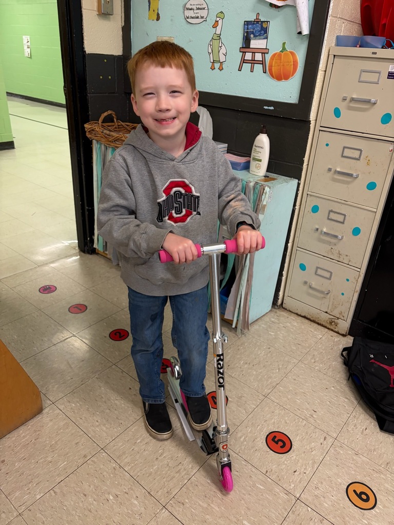 Photo taken inside a school hallway showing a student standing and holding a scooter. The student is wearing a hooded sweatshirt, jeans, and sneakers. The scooter has pink handle grips and wheels. Classroom items, including a filing cabinet, artwork on the wall, and floor markers, are visible in the background.