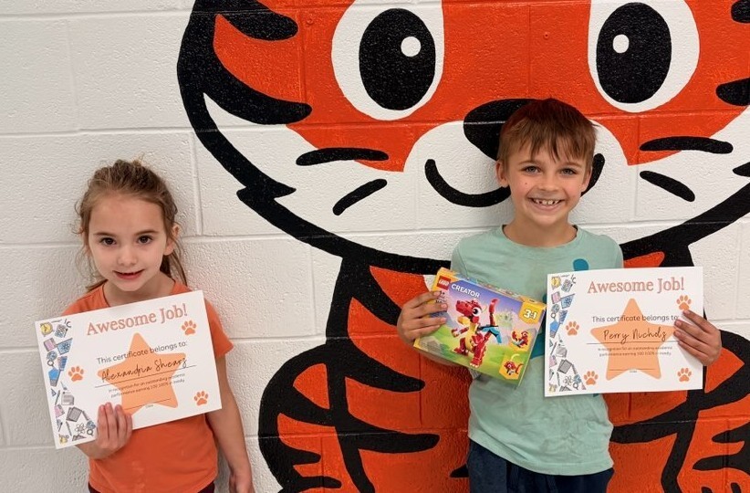 “Two students stand in front of a painted tiger mural on a cinderblock wall. Each student is holding an ‘Awesome Job!’ certificate with their name written on it. One student also holds a small prize.”