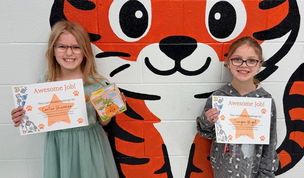 “Two students stand in front of a painted tiger mural on a cinderblock wall. Each student is holding an ‘Awesome Job!’ certificate with their name written on it. One student also holds a small prize.”