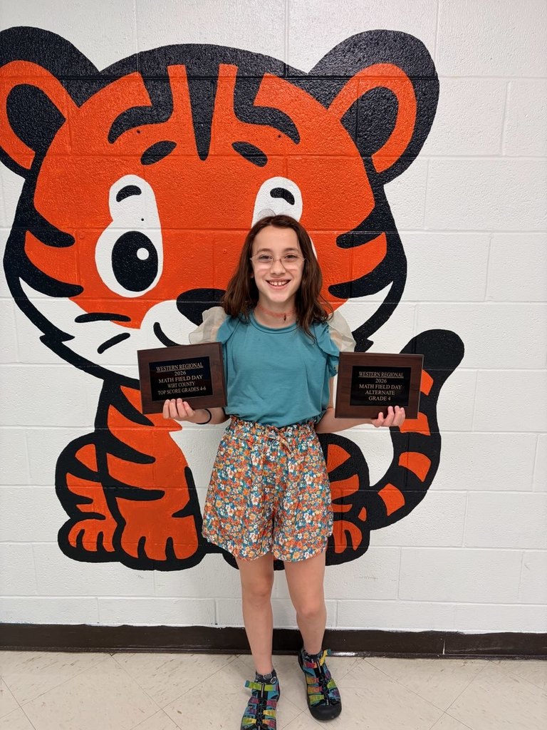 “A student stands in front of a painted tiger mural on a cinderblock wall while holding two rectangular math award plaques, one in each hand. The plaques recognize achievements in a regional math field day and a countywide math competition.”