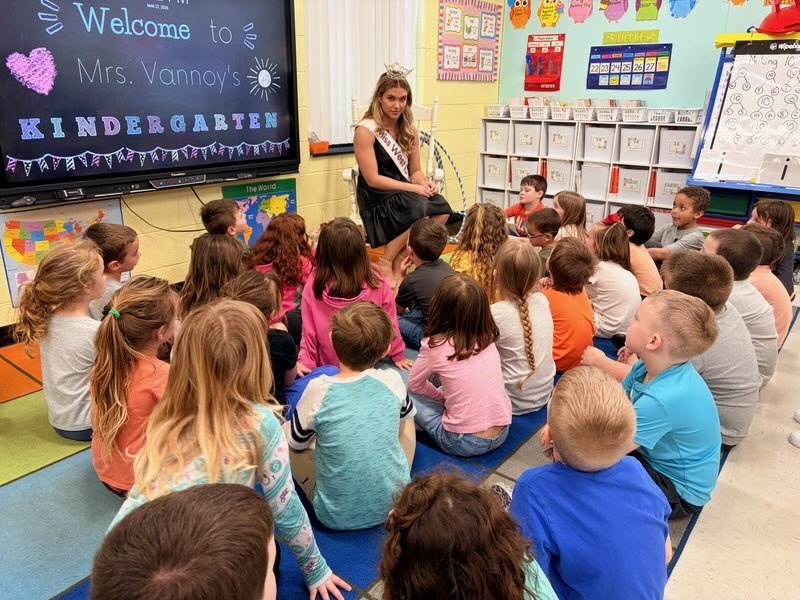“A classroom scene with multiple kindergarten students sitting on the floor and facing a seated guest wearing a sash and a crown. The guest is reading and speaking to the students. A classroom display board in the background reads, ‘Welcome to Mrs. Vannoy’s Kindergarten,’ and colorful posters and storage bins are visible around the room.”