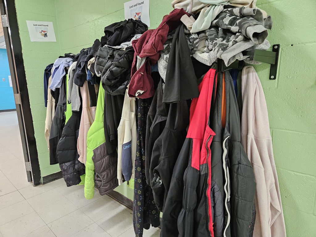 “Several jackets and hoodies of different colors and sizes hang closely together on a metal clothing rack in the school hallway.”