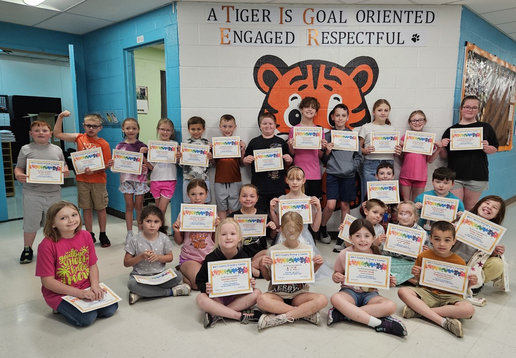 A group of elementary-aged students posed indoors in a school hallway, holding colorful certificates for perfect attendance. The students are arranged in several rows. Behind them is a wall mural featuring a large cartoon tiger and the words, “A Tiger Is Goal Oriented, Engaged, Respectful.”
