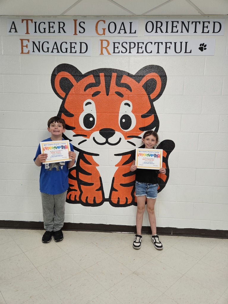 Two elementary-aged students stand in a school hallway holding perfect attendance certificates. They are positioned in front of a painted wall mural of a large cartoon tiger with the words, “A Tiger Is Goal Oriented, Engaged, Respectful,” displayed above the image.