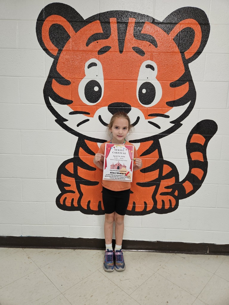 A student stands in a school hallway in front of a painted tiger mural, holding a Spring Carnival certificate that reads “Spring Carnival, April 24, 2025” with the name Alex Shears printed on it.