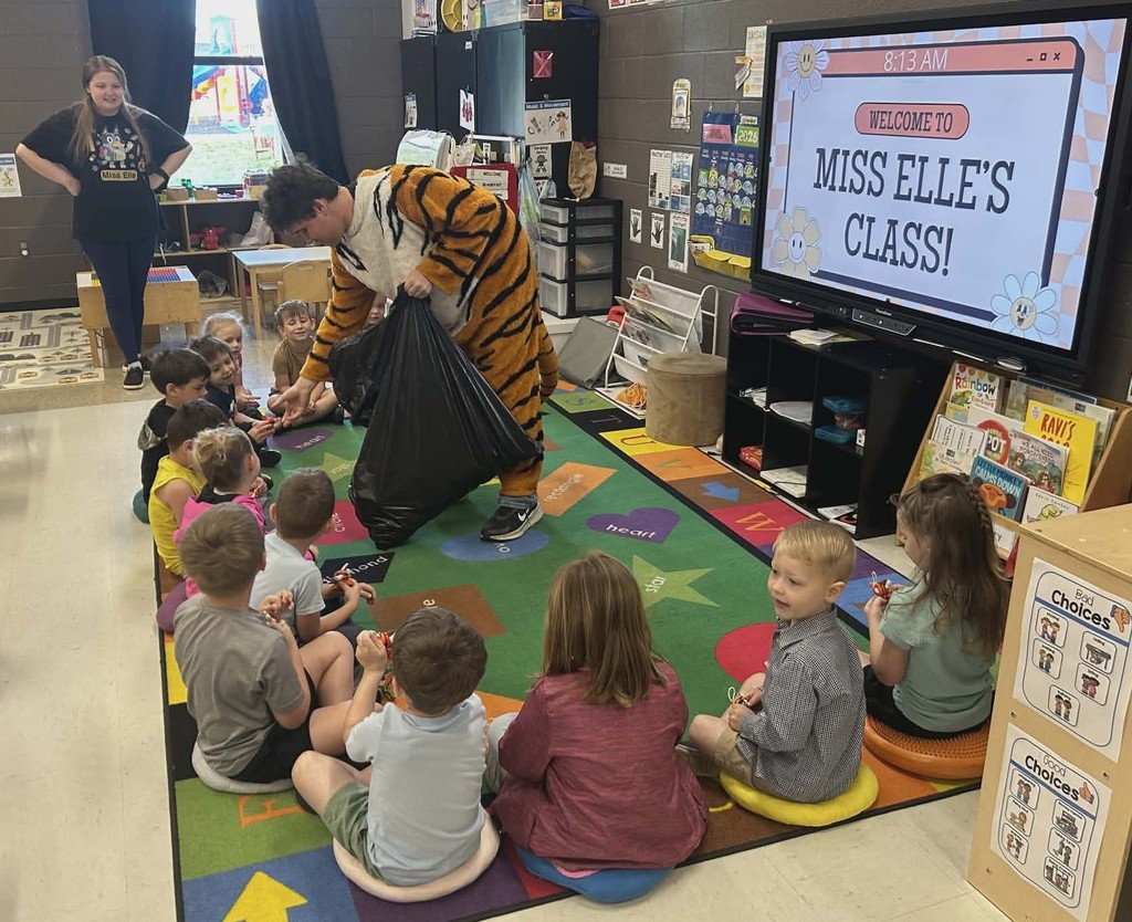 A school mascot dressed as a tiger stands inside an elementary classroom, handing out small stuffed tiger toys to a group of young students seated in a circle on a colorful classroom rug. Classroom shelves, books, and a smartboard that reads “Welcome to Miss Elle’s Class!” are visible in the background.