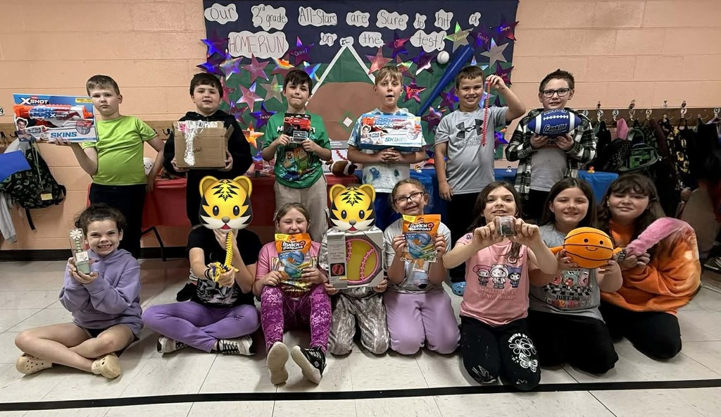 Group of third grade students posing indoors at school, arranged in two rows, holding various prizes such as toy sets, sports balls, games, and small boxes won from the Lucky Duck attendance contest. A decorated bulletin board is displayed behind the students.