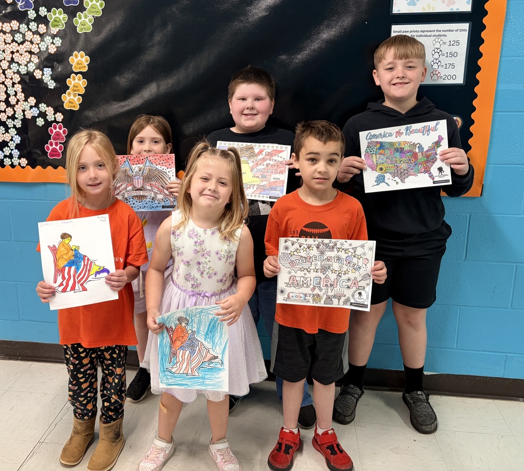 A group of five elementary-aged students from Wirt County Primary Center stand in a school hallway in front of a black bulletin board and blue wall, each holding a colored drawing from a patriotic-themed coloring contest. The drawings include images such as a map of the United States labeled “America the Beautiful,” an eagle design with “Since 1776,” and scenes related to American history. The students are arranged in a row, with some standing slightly in front of others. A display of colorful paw print cutouts and a classroom poster are visible in the background.