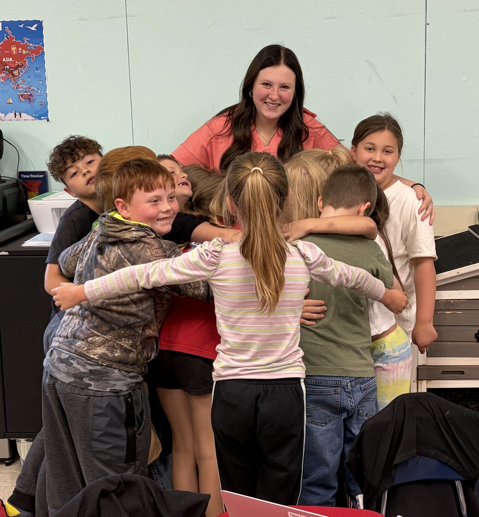A group of elementary students in a classroom gather around and hug senior intern Carly Phillips as they say goodbye. Carly stands smiling in the center while students surround her with their arms around one another, showing affection and appreciation. The classroom walls and furniture are visible in the background.
