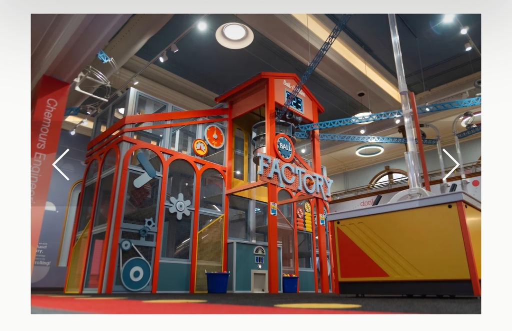 Colorful indoor children’s science play area with a large red and orange structure labeled “Factory,” featuring gears, tubes, and interactive elements under bright overhead lights, representing a hands-on learning exhibit at a discovery museum.