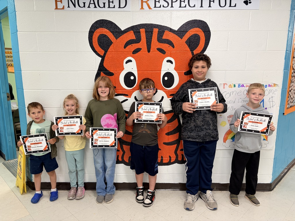 Six elementary-aged students stand side by side in a school hallway, each holding a “Student of the Month – April 2026” certificate. Behind them is a painted mural of a large orange-and-black tiger face on a white brick wall. The students are positioned in front of the mural and spaced evenly across the frame. A colorful poster board is visible on the right side of the image, and a doorway with blue trim appears on the left. The students are wearing casual clothing and facing the camera while displaying their awards.