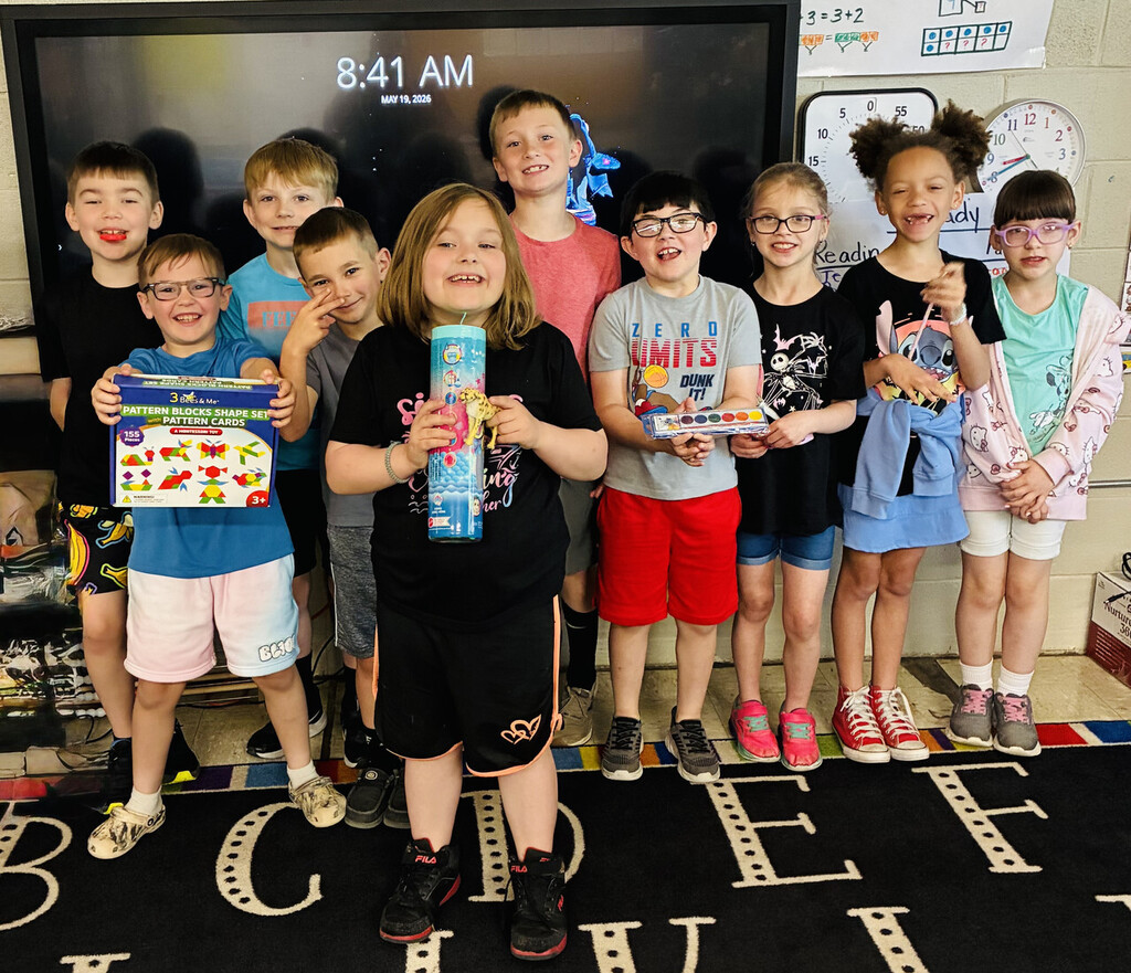 Ms. Lowe's students stand together in a classroom in front of a large digital display that shows the time and date. The students are arranged in a row and are holding various prize items, including a box of pattern blocks, a tall cylindrical toy container, and a watercolor paint set. A classroom wall behind them includes educational posters and an analog clock. The students are standing on a rug with large letters and are facing the camera.