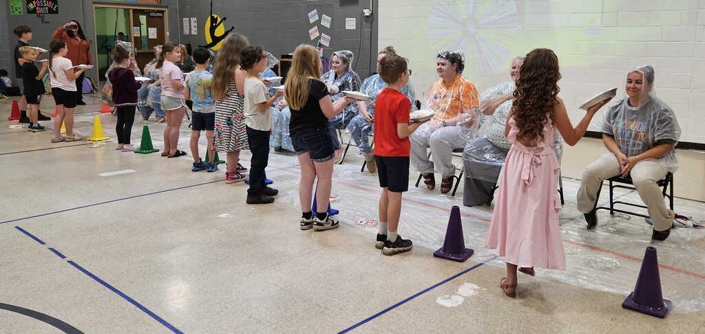 Students stand in a line inside a school gym holding whipped-cream pies while several teachers and aides sit in chairs wearing plastic ponchos and hair coverings. On a signal, the students press the pies into the adults’ faces at the same time, creating a messy scene with cream on faces, clothing, and the floor. Colored cones and a projected spinning wheel are visible in the background.