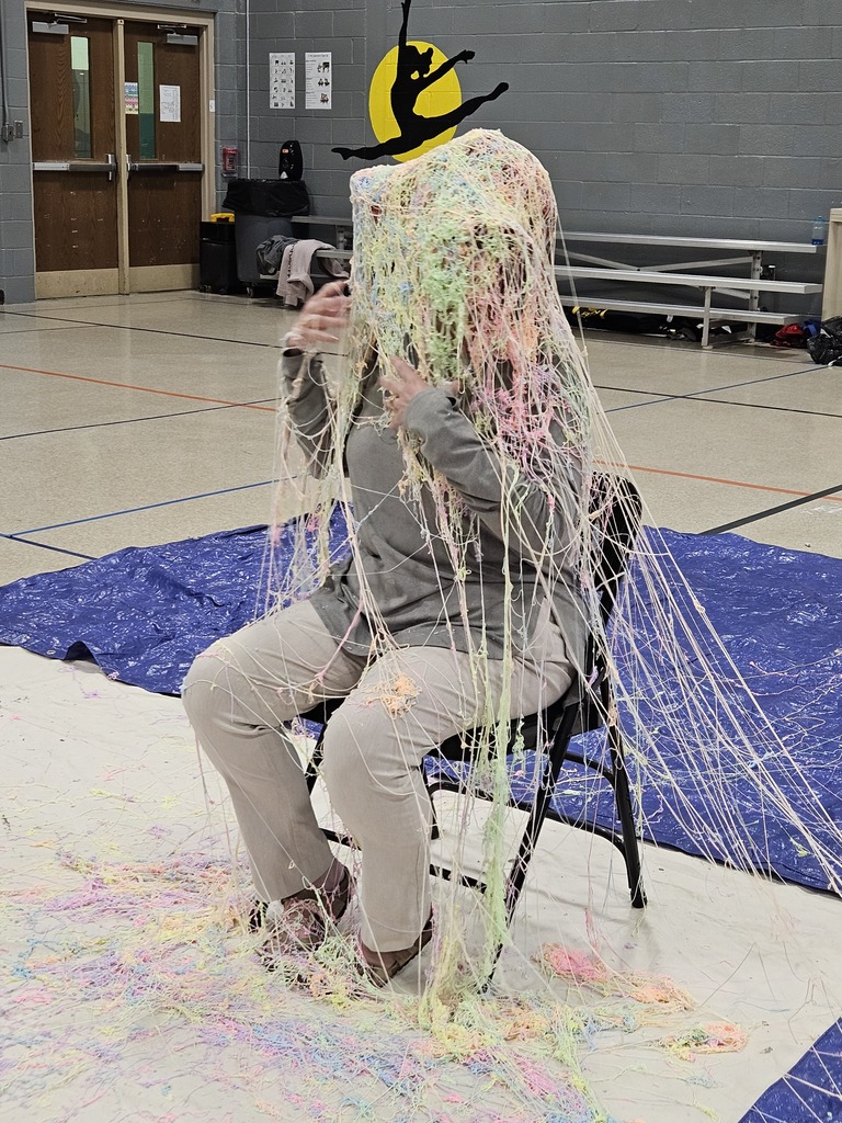 A person sits on a chair in a school gymnasium, covered from head to knees in long strands of colorful Silly String in shades of pink, yellow, green, and white. The string drapes over the person’s clothing and pools on the floor and a blue tarp underneath. Gym walls, double doors, and bleachers are visible in the background.