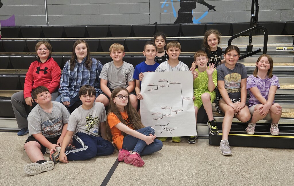 A group of approximately 14 elementary students sit and kneel together on indoor gym bleachers at Wirt County Primary Center. The students are arranged in two rows, with some sitting on the floor in front and others seated on the bleachers behind. One student in the center holds a large poster board displaying a hand-drawn tournament bracket with team names and scores from a wiffle ball competition. The group is posed closely together in a gym setting with black bleachers and a painted wall in the background.