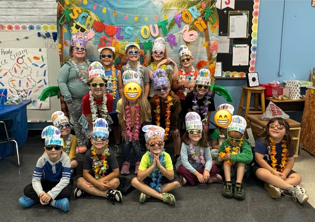 A group of elementary students pose together in a decorated classroom for a “Beach Bash” celebration. The students are wearing colorful leis, sunglasses, and paper hats with drawings and writing. They are arranged in rows in front of a tropical-themed backdrop with bright decorations and a banner. Some students are standing while others sit on the floor. Classroom items such as a whiteboard, posters, and supplies are visible around the room.