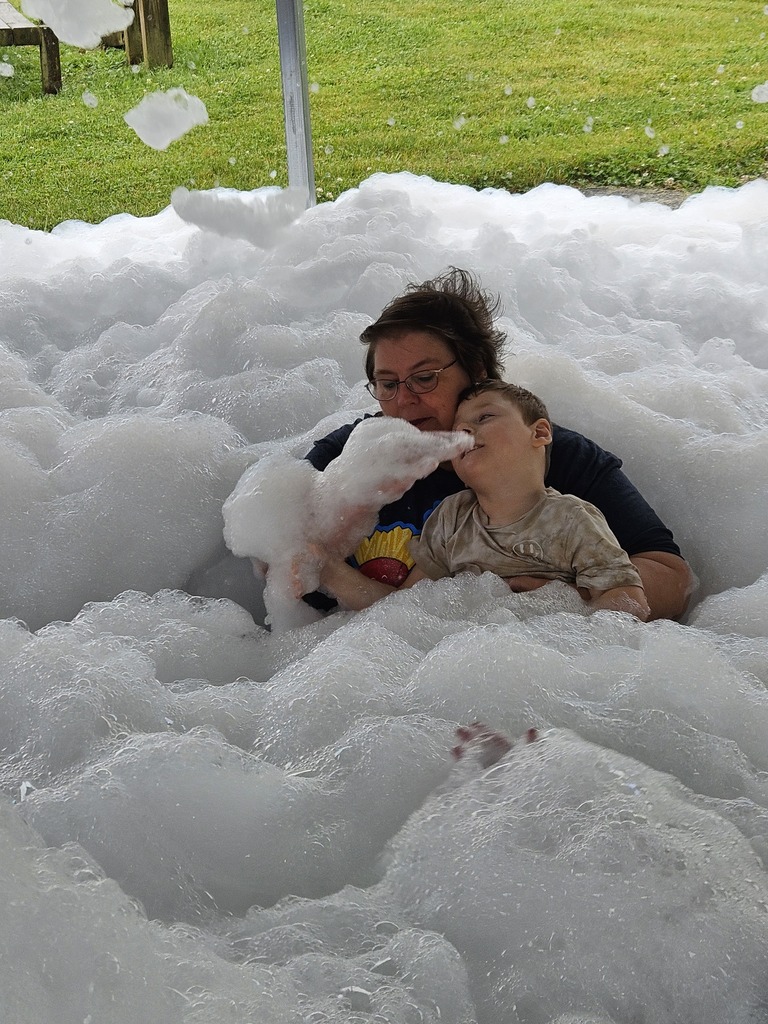An adult and a young student sit together under a covered outdoor area surrounded by a large pile of white foam during a school foam party. The two are partially covered in bubbles, with foam piled around them and floating through the air, while green grass is visible beyond the shelter in the background. 