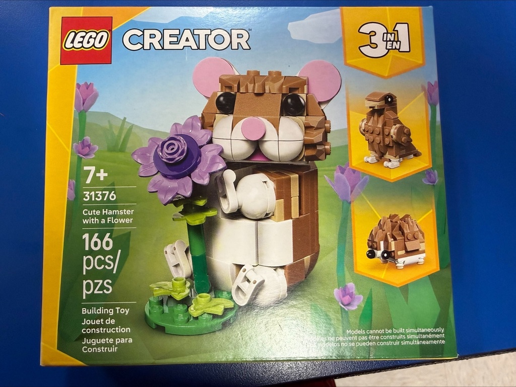 Photograph of a LEGO Creator 3-in-1 building set box displayed on a blue surface. The set is LEGO Creator model 31376, "Cute Hamster with a Flower," recommended for ages 7 and up and containing 166 pieces. The front of the box shows a brown and white hamster holding a purple flower in a grassy setting with flowers in the background. Images on the right side of the box show two alternate builds: a small bird and a hedgehog. The LEGO logo and "Creator 3 in 1" branding are visible on the packaging.