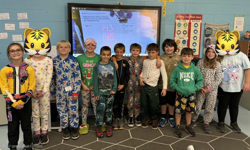 A group of elementary students from Mrs. Roberts' class stands together in a classroom for Pajama Day during Homecoming Spirit Week. The students are wearing a variety of pajamas, including colorful pajama sets, character-themed sleepwear, and comfortable clothing. An interactive classroom display screen and educational posters are visible in the background. The class is gathered side by side and posing for a group photo.