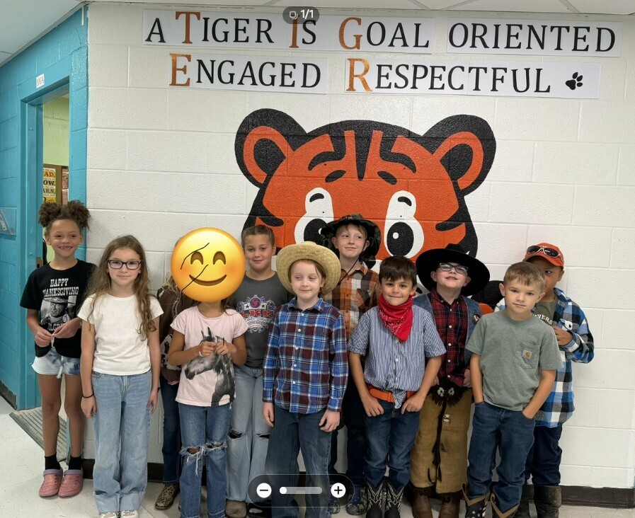 A group of elementary school students from Mrs. Carouther's class stand in front of a large tiger mural and a school display that reads "A TIGER is Goal Oriented, Engaged, Respectful." Several students are dressed for Western Day during Spirit Week, wearing cowboy hats, plaid shirts, bandanas, boots, and other western-themed clothing. The students are posing together in a school hallway for a class photo.