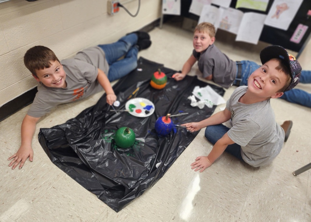 Photos show elementary students participating in a PBIS reward activity by painting pumpkins in various rooms throughout the school. Students use paint and art supplies to decorate pumpkins in different colors and designs while working at tables and classroom spaces. The pumpkins were provided by Moore’s Piggly Wiggly for students who earned enough PBIS points to participate in the fall activity.