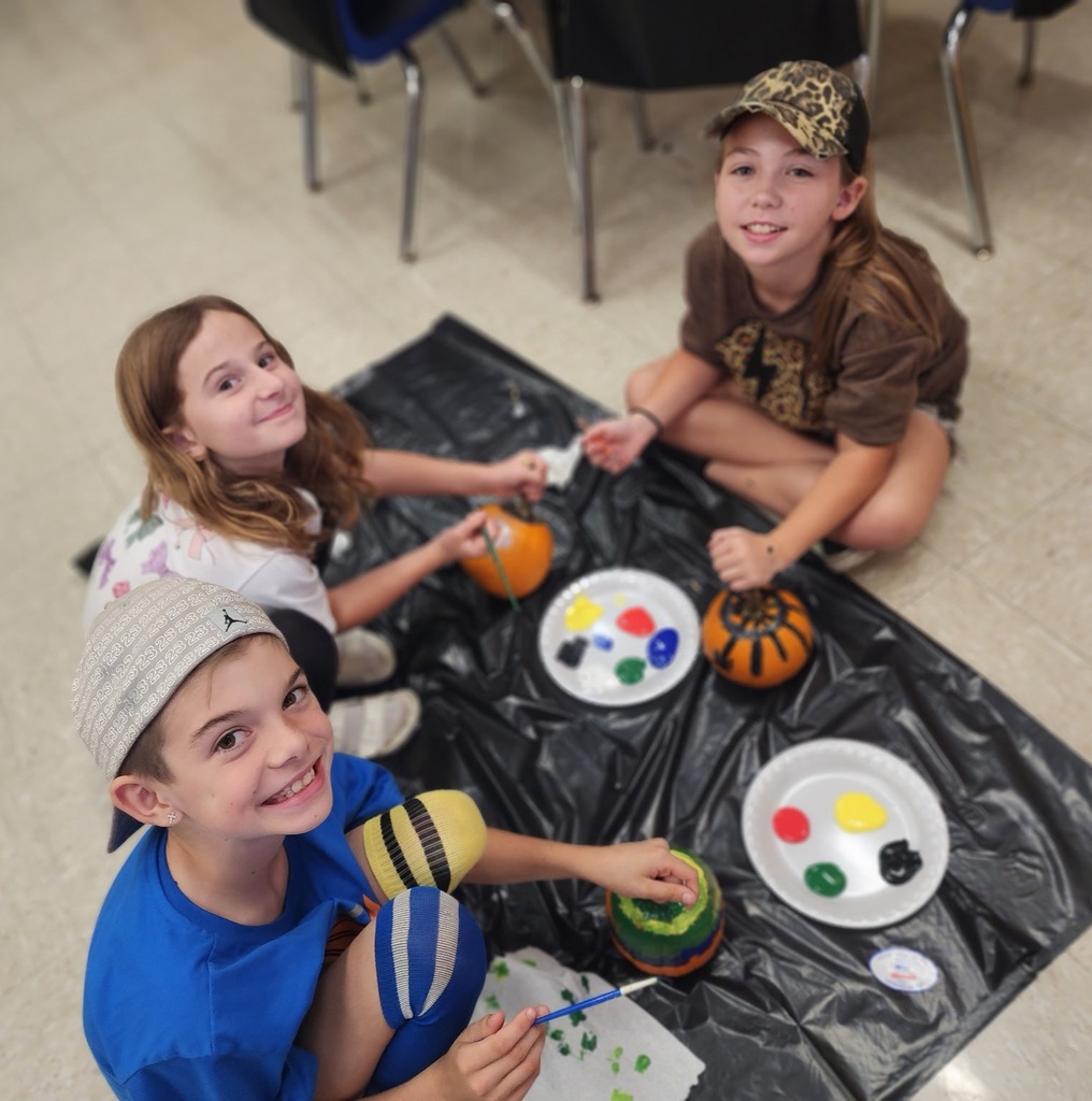 Photos show elementary students participating in a PBIS reward activity by painting pumpkins in various rooms throughout the school. Students use paint and art supplies to decorate pumpkins in different colors and designs while working at tables and classroom spaces. The pumpkins were provided by Moore’s Piggly Wiggly for students who earned enough PBIS points to participate in the fall activity.